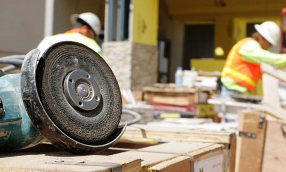 A close-up of a power grinder on a construction site, with workers in safety vests and helmets in the background.