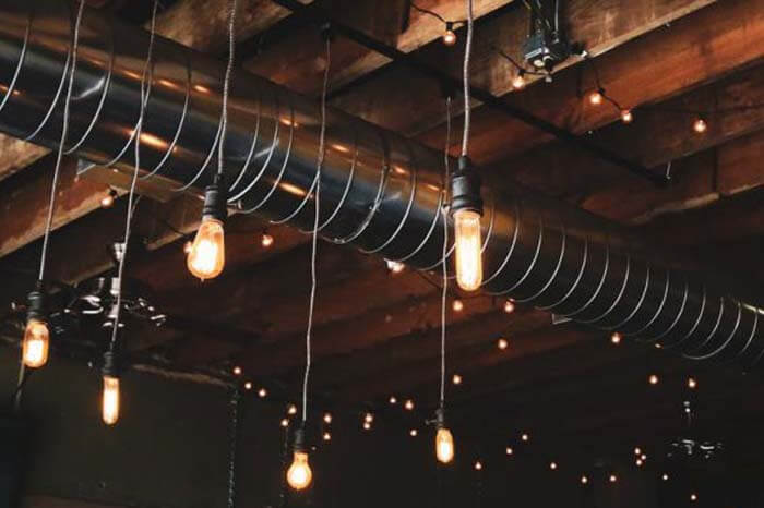 Islington Electrical Industrial-style ceiling with exposed wooden beams, metal ventilation ductwork, and hanging light bulbs.